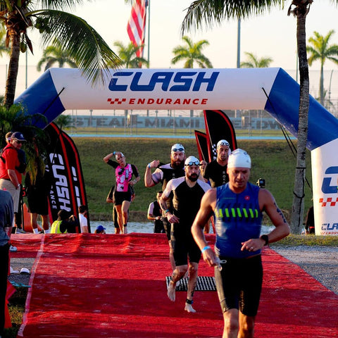 Triathletes pass under a sturdy PVC inflatable arch emblazoned with CLASH Endurance at a race event, highlighted by the morning sun and swaying palm trees.