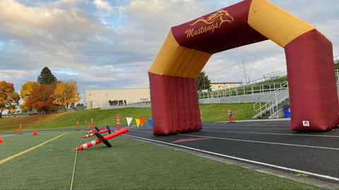 A maroon and gold denier polyester inflatable arch, proudly displaying Mustangs marks the start of a race at Milwaukie High School against a backdrop of autumn trees.