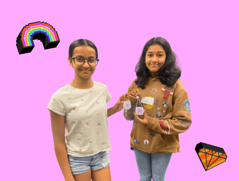 Two middle school girls stand holding their imagi charms proudly in front of a bright pink background.