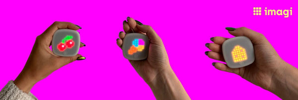 3 hands hold colorful imagiCharms displaying pixel art against a pink background