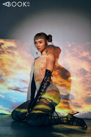 Influencer Lil Miquela kneeling in front of a sky background wearing a peach colored dress with black latex gloves and high heels