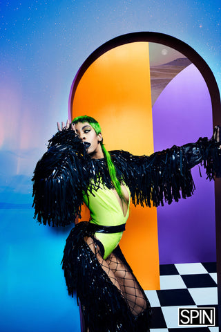 Rico nasty wearing neon green deep v Vex body suit with mesh tights, matching black fringed chaps and bolero with green hair for a photoshoot posing again multicolored walls