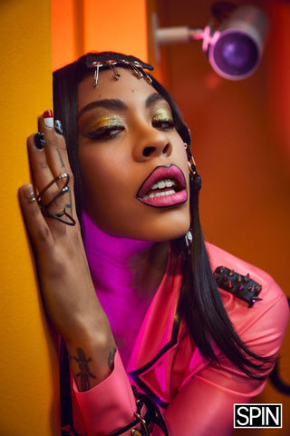 Rico Nasty wearing Vex custom pink spike trench for a photoshoot posing close up with full makeup and pink lips against an orange wall
