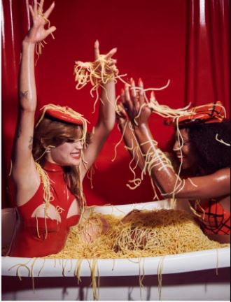 Two models in a bathtub of spaghetti wearing a red Vex mini beret and red divide bodysuit with a red backdrop