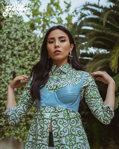 Woman wearing patterned top with blue bra over it posing in front of greenery