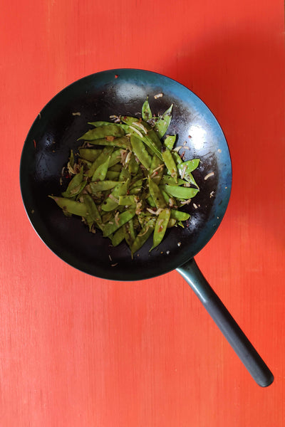 A carbon steel wok with green snow peas, shallots, garlic and chili on a red background.