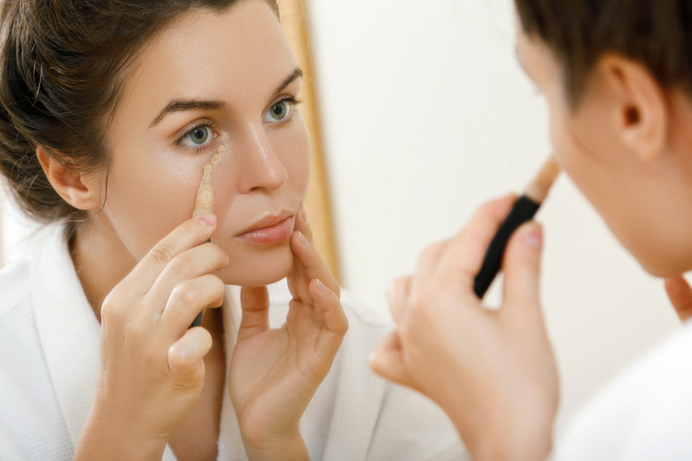 Woman looking in the mirror while applying concealer under her eyes.