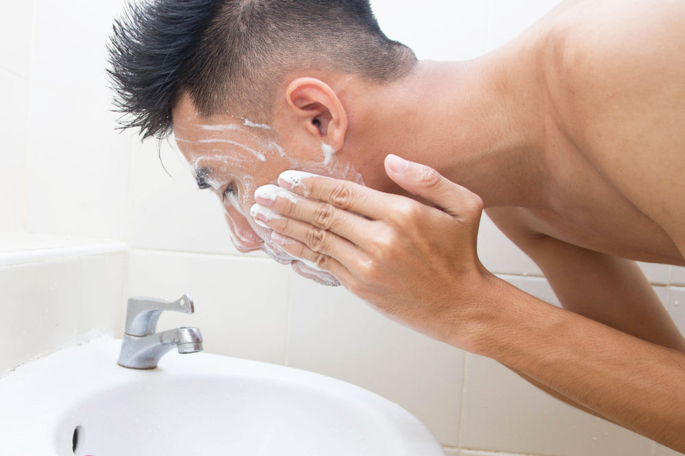 Asian teen boy washing his face over a white sink.