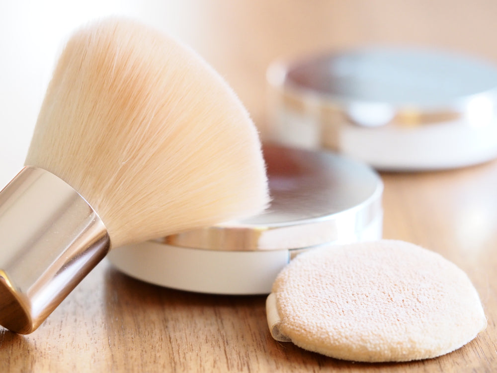 Kabuki brush, powder compact, and makeup round sitting on a wood surface.