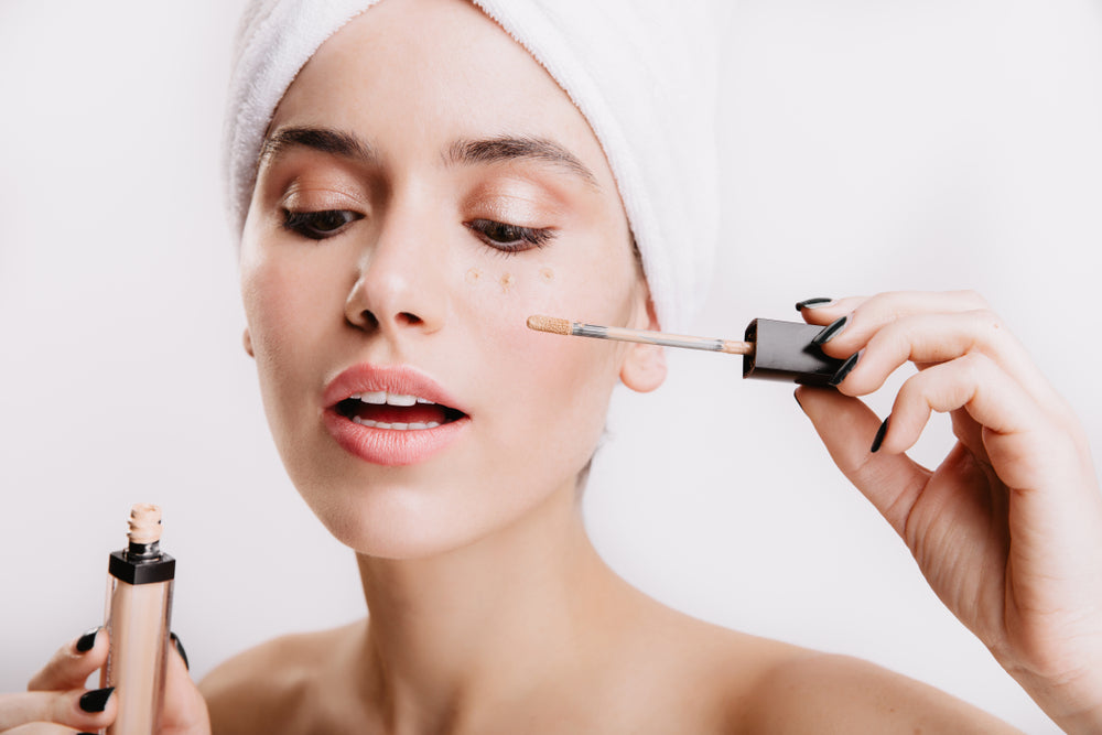 Woman wearing a hair towel holding a concealer wand up to her face.