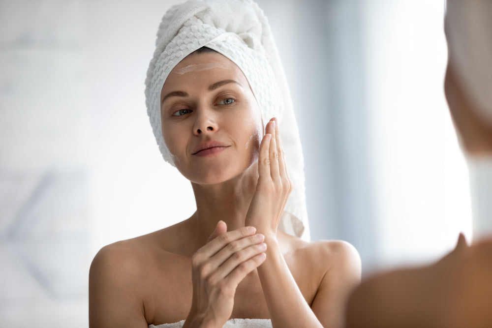 Woman standing in front of the mirror in a towel applying sunscreen to her face.