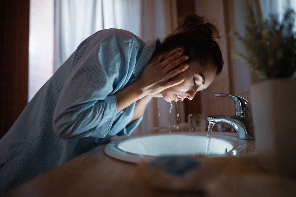Image of brunette woman washing her face in the bathroom at night.