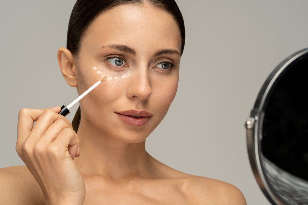 Image of a woman looking in the mirror while applying concealer under her eye.