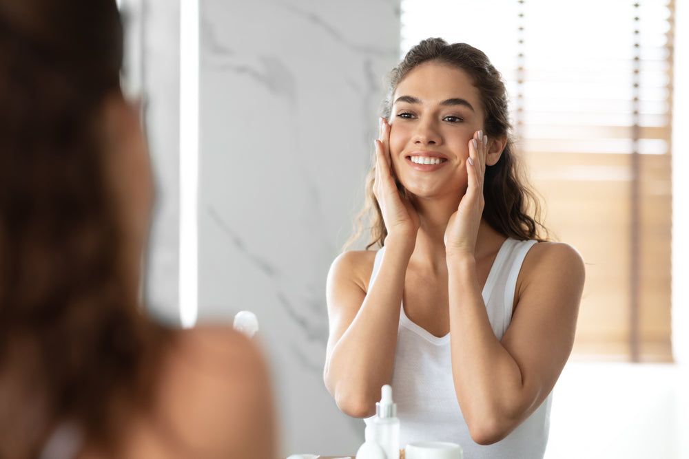 White woman looking at herself in the mirror smiling and cupping her cheeks.