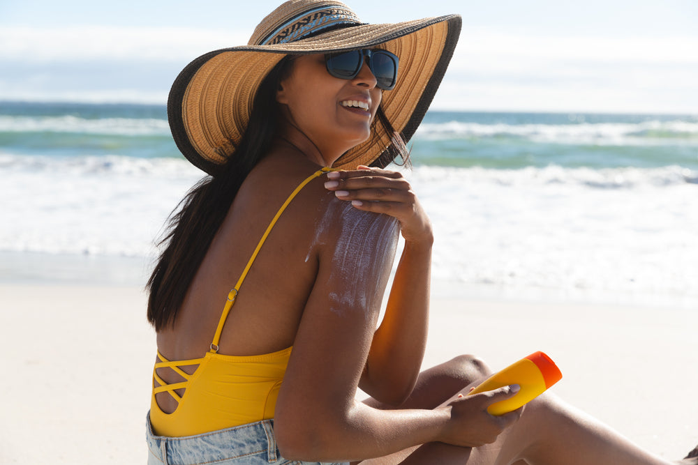 Woman in a yellow bathing suit and sun hat sitting on the shore applying sunscreen lotion.