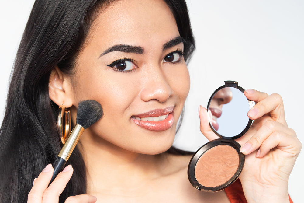 Woman putting bronzer makeup on her face while holding up her handheld mirror