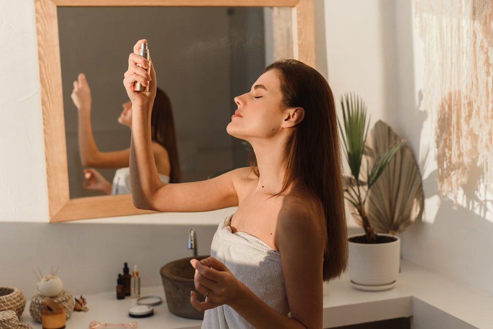 Woman standing in bathroom in her towel applying setting spray to her face.