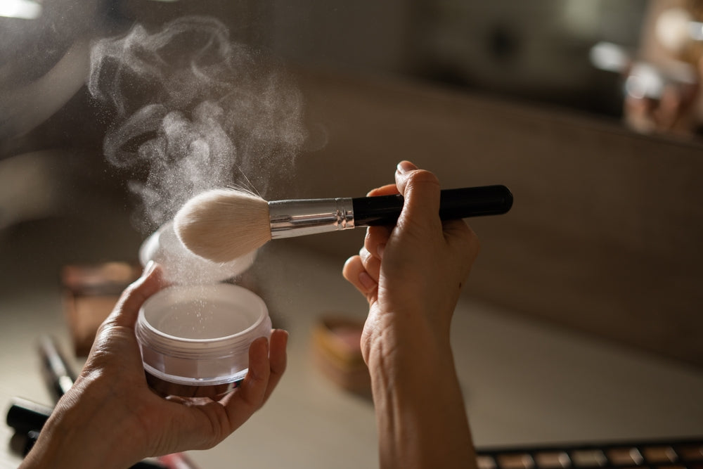 Close up of a hand holding a setting powder container and the other holding a brush with powder drifting in the air.