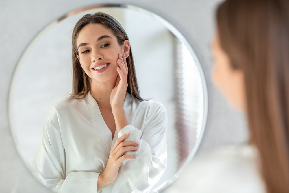 Brunette woman in a robe smiling and looking at her reflection in a circular mirror.