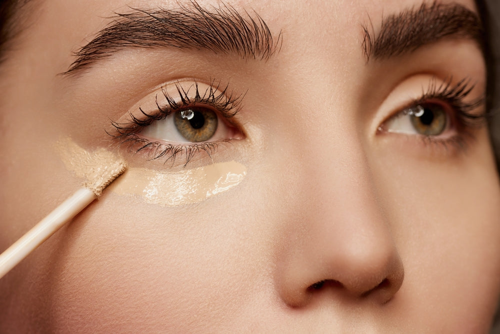 Close up of a woman’s face, showing her applying concealer to her under eye.