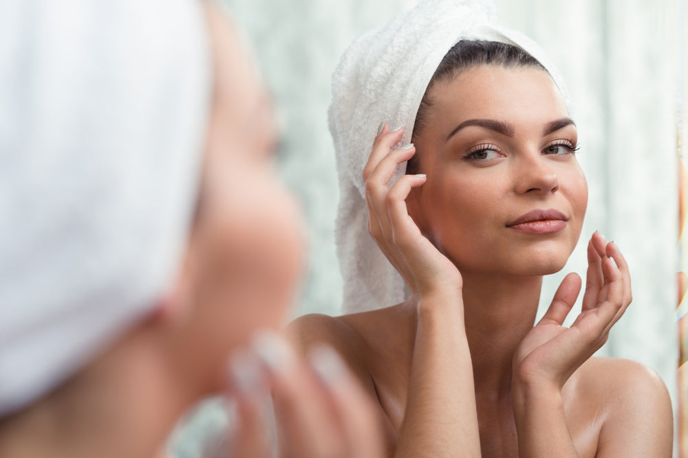 Woman looking at her skin in the mirror with her hair in a towel.