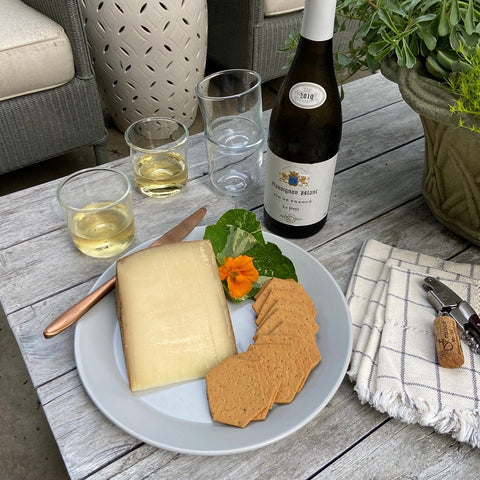 Dinner plate with wedge of cheese, crackers, and orange flower next to white wine in glasses and stack of white cloth napkins