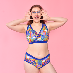 Alice is wearing a retro rainbows bra and briefs against a pink background.  She has her hands to her face in a 'wow' expression