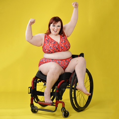Model is sitting jauntily in a wheelchair with her hands in the air. She's wearing a berries and cherries print underwear set which is mostly red.