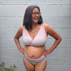 Model is standing outside in front of a concrete wall. She is wearing a grey and pink underwear set.
