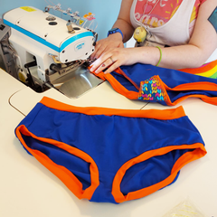 The hands of a machinist at their machine, sewing the leg of a pair of blue and orange pants with a knit gusset. A completed pair is laid out in front of them.