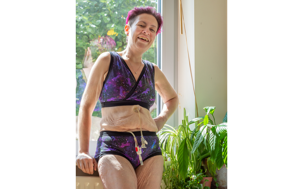 Imogen is standing at a window resting on the sill with one hand. They are wearing a Uni-verse underwear set and have some plants on their right.