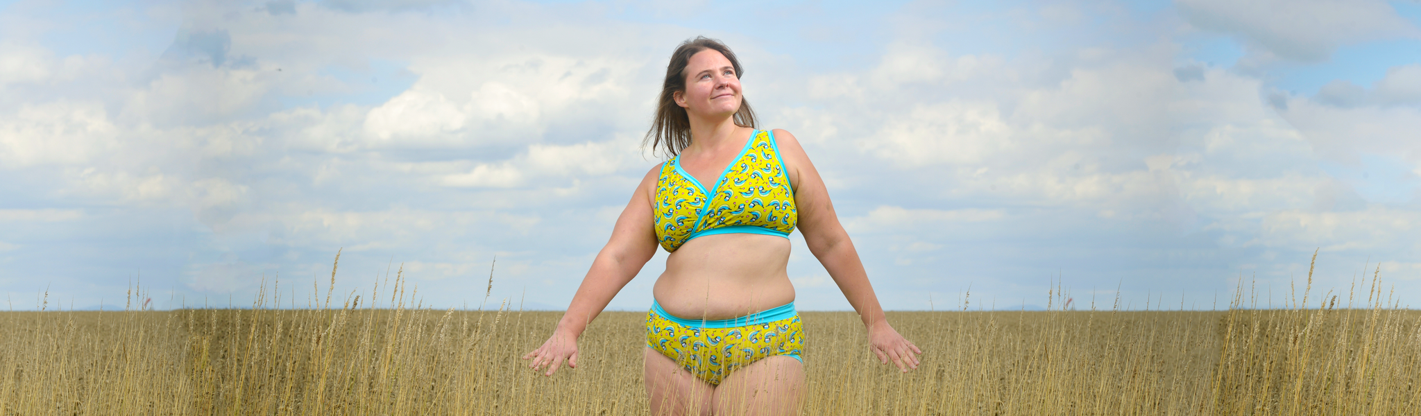 A woman stands in a field of wheat with her arms out and the sky is blue. She looks content. She is wearing a set of Molke underwear in the Blue Tits fabric. It is yellow with illustrated birds on it an a light blue trim.