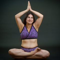 Model is sitting down in a yoga pose with her palms together abover her head. She is wearing a purple lace print underwear set