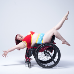 Model is sitting in a wheelchair backwards leaning back with legs and arms outstretched. She is wearing a Power underwear set which is a red bra and blue pants with stars on them.