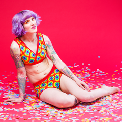 Model is sitting on the ground surrounded by confetti. She is wearing a rainbow circles underwear set and has purple hair and tattoos.