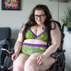 Sarah sat in her wheelchair in her home smiling softly. She is wearing the Dino set which is green with small dinosaurs on it and a purple trim.