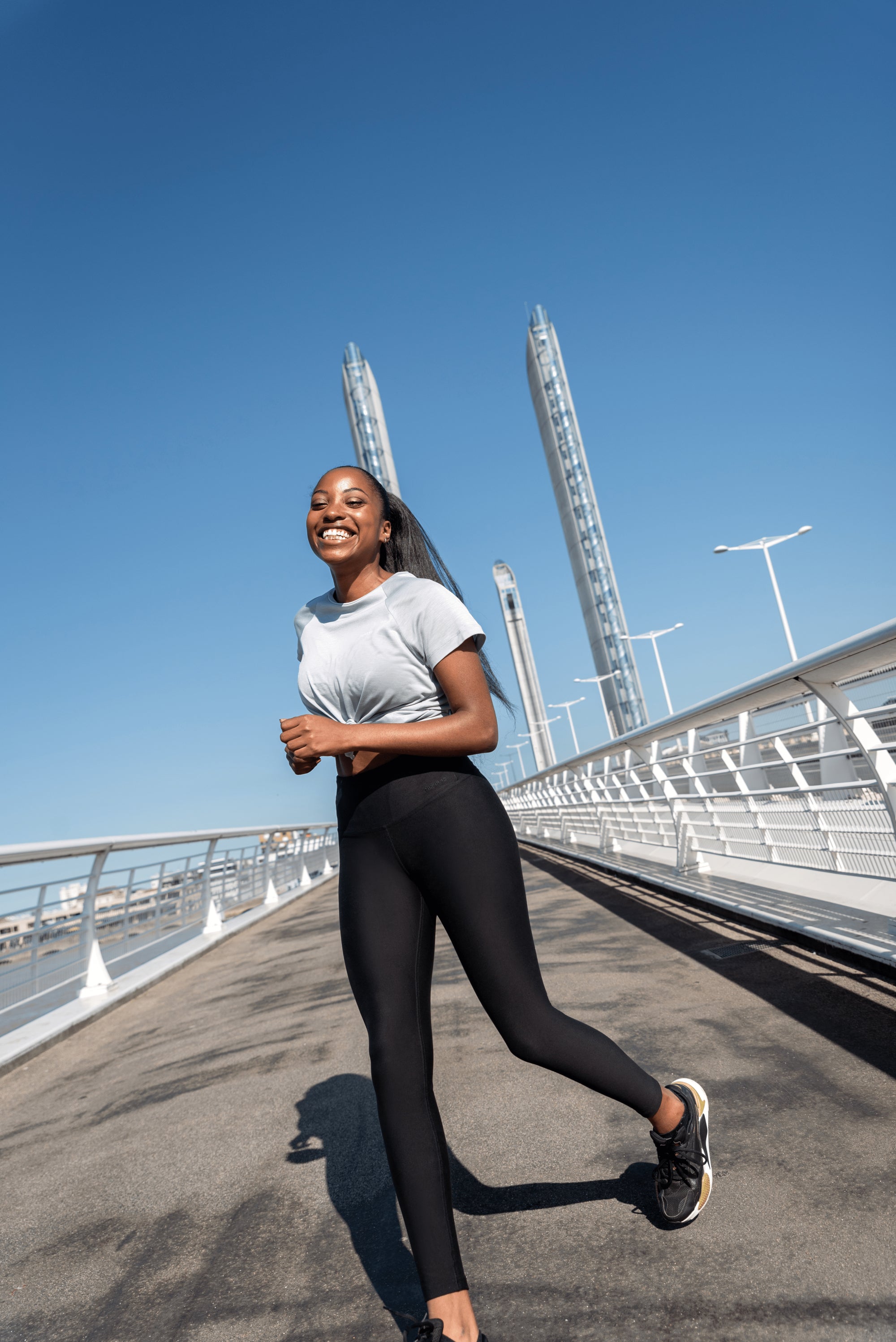 Woman with dark hair and complexion running outside on a pedestrian bridge in the sunshine. Woman is wearing Tripulse Original Workout T-Shirt in colour a timeless light grey colour. She is also wearing Tripulse Original Leggings 2.0 in classic black.
