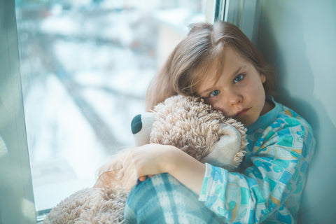 Young girl cuddling a teddy bear in a window in her pajamas looking a little unwell.