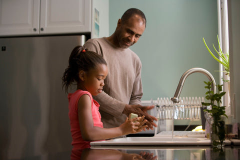 Dark skinned father with short hair watches over his young daughter, also dark skinned with a light red singlet top on ona a ponytail in her hair wash her hands with a bar of soap over the kitchen sink.