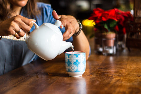 A white skinned lady pours a cup of tea in a handleless mug with blue diamonds on it from a white teapot.