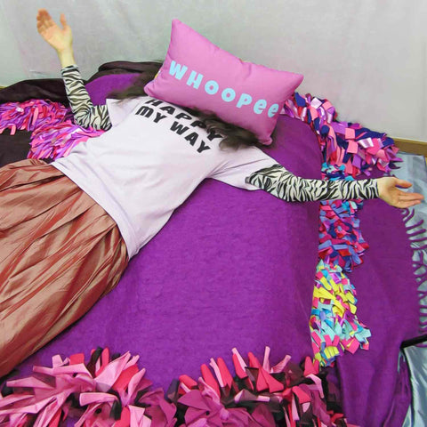 Alex Mitchell reclining in colorful setting with a Whoopee slogan pillow covering her face.