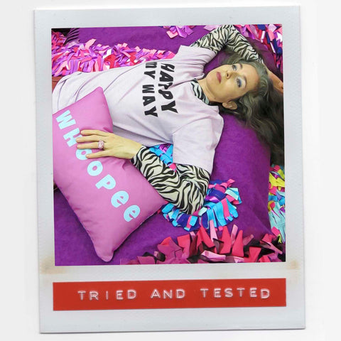 Alex Mitchell reclining in colorful setting wearing Happy My Way slogan tee surrounded by boa scarves.