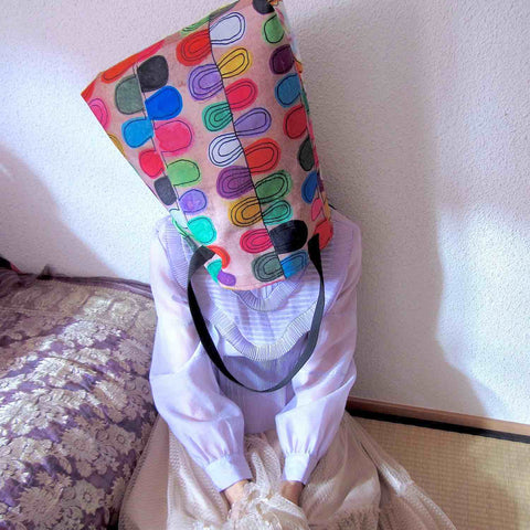 Alex Mitchell in a silly sitting pose wearing a big colorful tote bag in Frosted Cookies-Flipflop design on her head for a reset break.