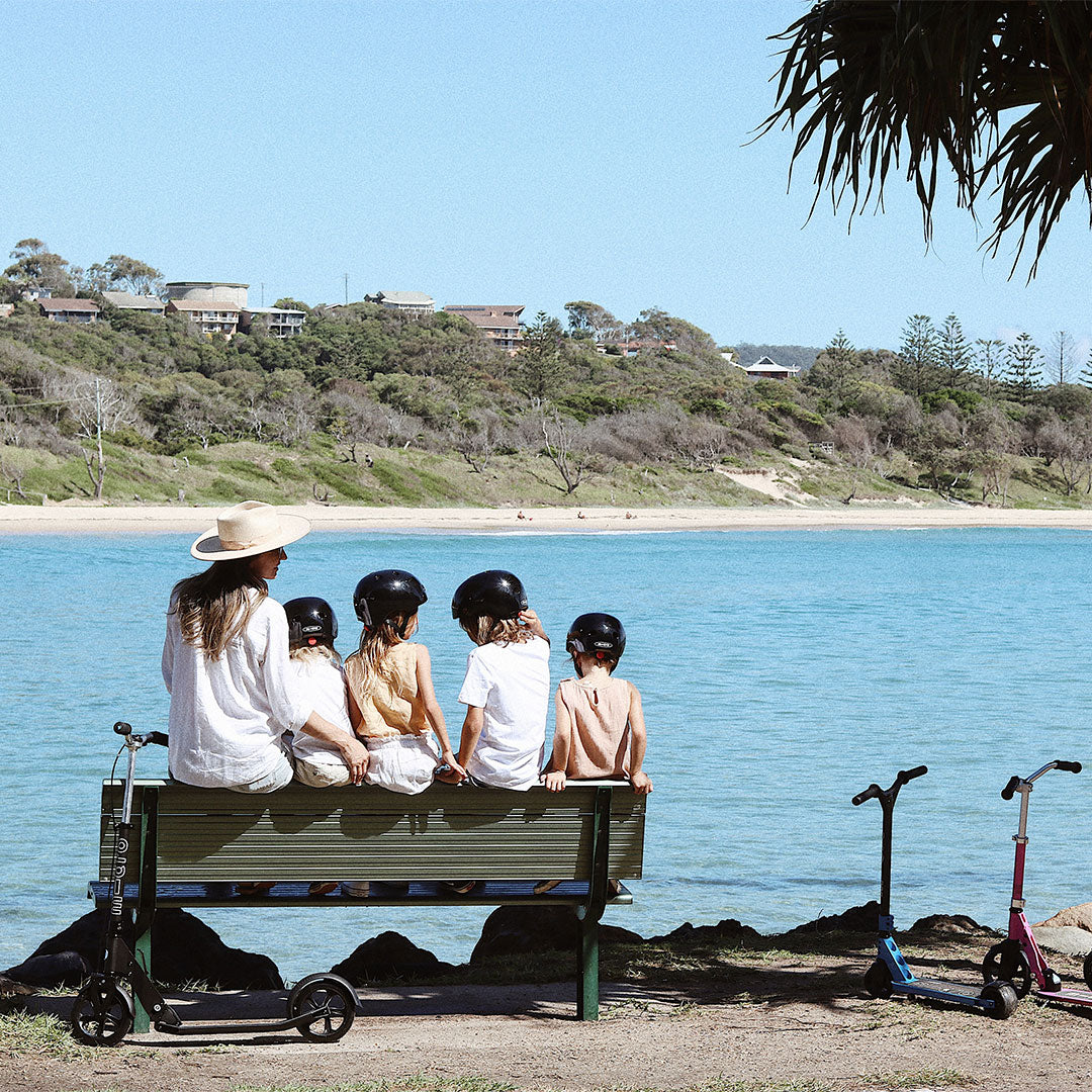 Mum and children sitting on a park bench with their scooters looking at the water