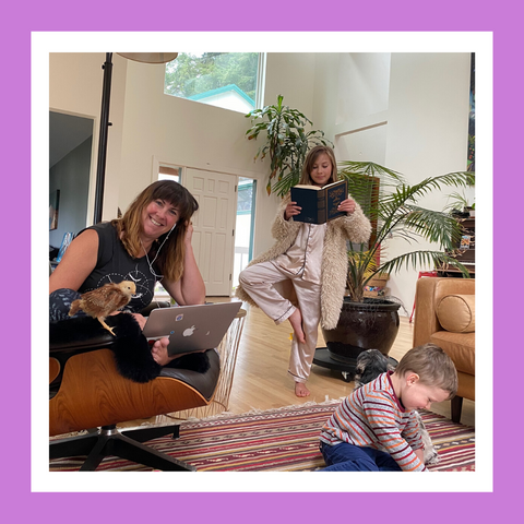 Woman wearing headphones working on a laptop with a girl doing a yoga pose while reading a book and a child on the floor