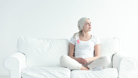 Woman undergoing cancer treatment sitting on a white couch in white clothes wearing pink cancer pin - looking towards side of image