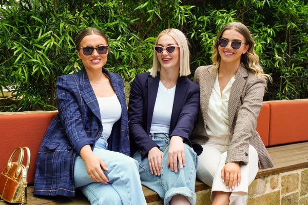 Belle, Jasmine and Jordei seated together outdoors, smiling and wearing trendy Glamour Glasses sunglasses, with a lush green backdrop.