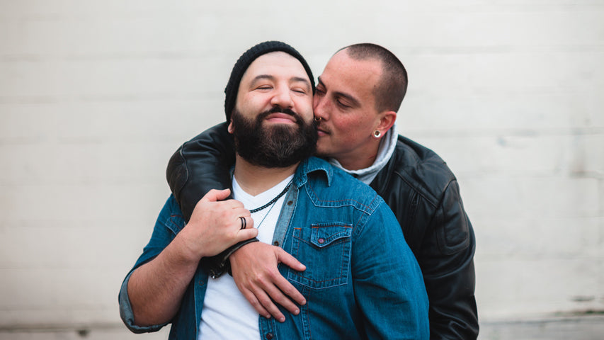 Man holding onto his partner while kissing his beard as his partner smiles against a white brick wall.