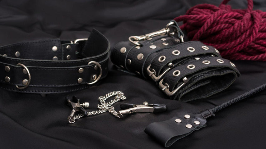 Des boucles de cuir et d’autres objets variés liés au BDSM sur un drap de lit noir.