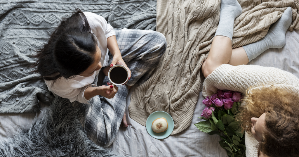 Woman holding tea while her girlfriend is clutching a bouquet of flowers atop blankets.